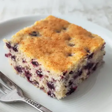 Ein Stück veganer Heidelbeer-Blechkuchen auf einem Teller, daneben eine Kuchengabel.