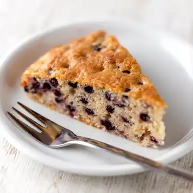 Ein Kuchenstück von einem veganem Heidelbeerkuchen auf einem weißen Teller, daneben eine Kuchengabel.