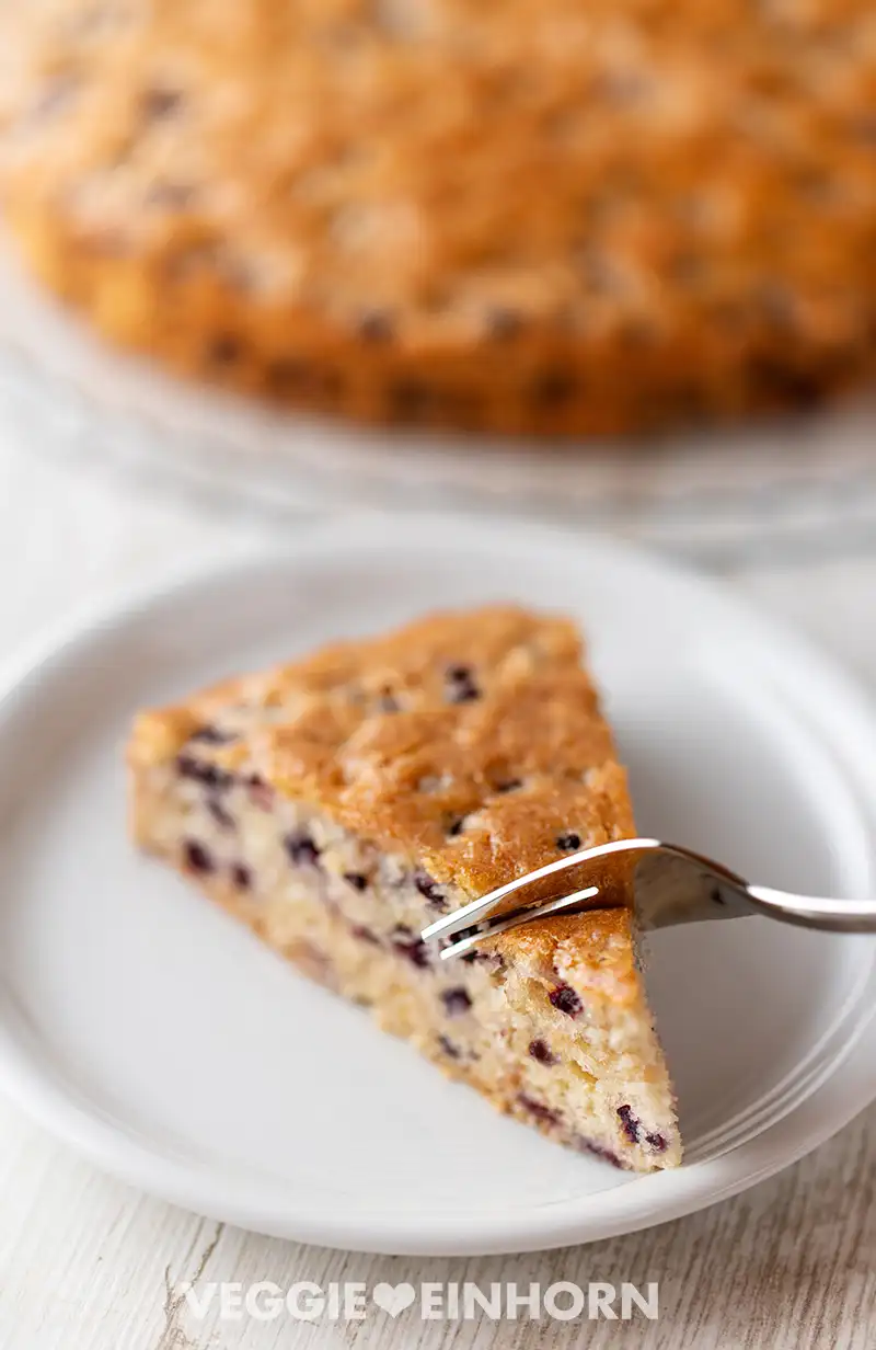 Kuchenstück mit Gabel auf einem Teller, im Hintergrund der restliche vegane Heidelbeerkuchen auf einer Tortenplatte.