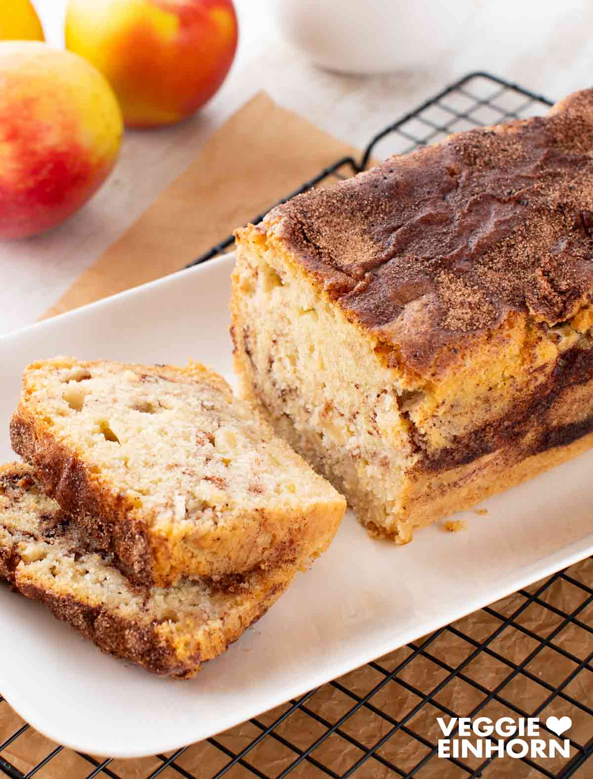 Ein veganer Zimtkuchen mit Apfel auf einer weißen Platte, teilweise aufgeschnitten. Im Hintergrund sind frische Äpfel, Backpapier und ein Kuchengitter zu sehen.
