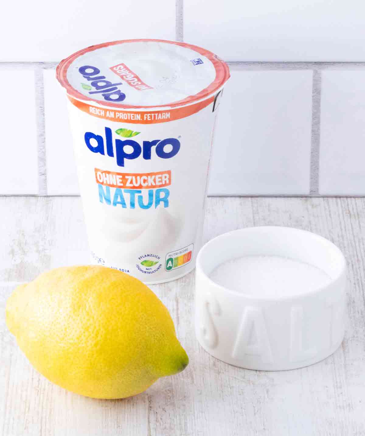Eine Zitrone, ein Becher Sojajoghurt ohne Zucker und Salz auf einem weißen Holzuntergrund.
