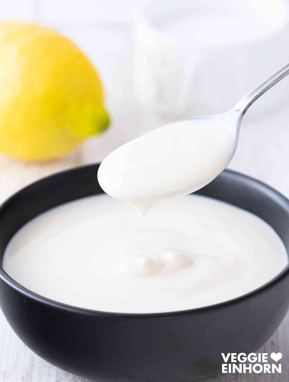 Löffel mit veganer Joghurtsauce über einer schwarzen Schale mit veganer Joghurtsauce.
