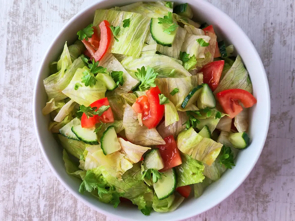 Eisbergsalat mit Tomaten, Gurken und Petersilie in einer weißen Schüssel auf einem hellen Holztisch.