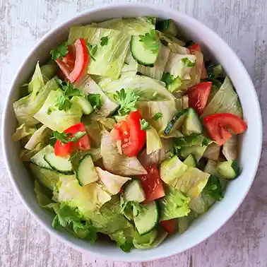 Ein weißer Teller mit frischem Eisbergsalat, Tomatenstücken, Gurkenscheiben und Petersilie auf einem hellen Holztisch.