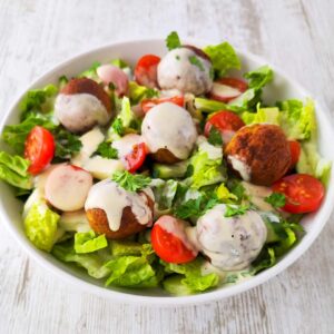 Falafel-Salat mit knusprigen Falafeln, Romanasalat, Kirschtomaten, Gurken und cremigem Dressing, serviert in einer weißen Schüssel auf einem hellen Holztisch.