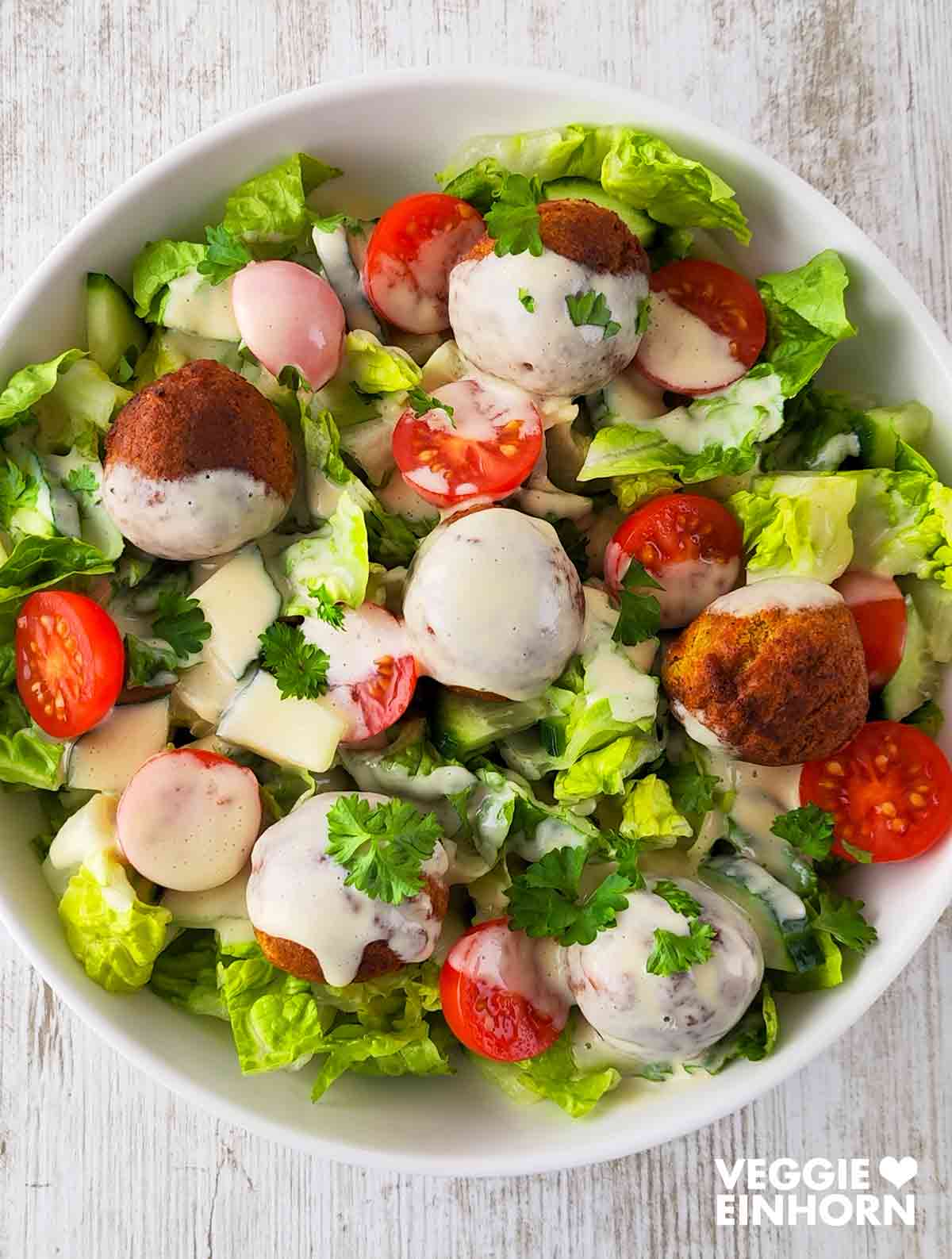 Bunter Falafel-Salat mit Romana Salatherzen, Kirschtomaten, Salatgurken und cremigem Dressing, serviert in einer weißen Schüssel.