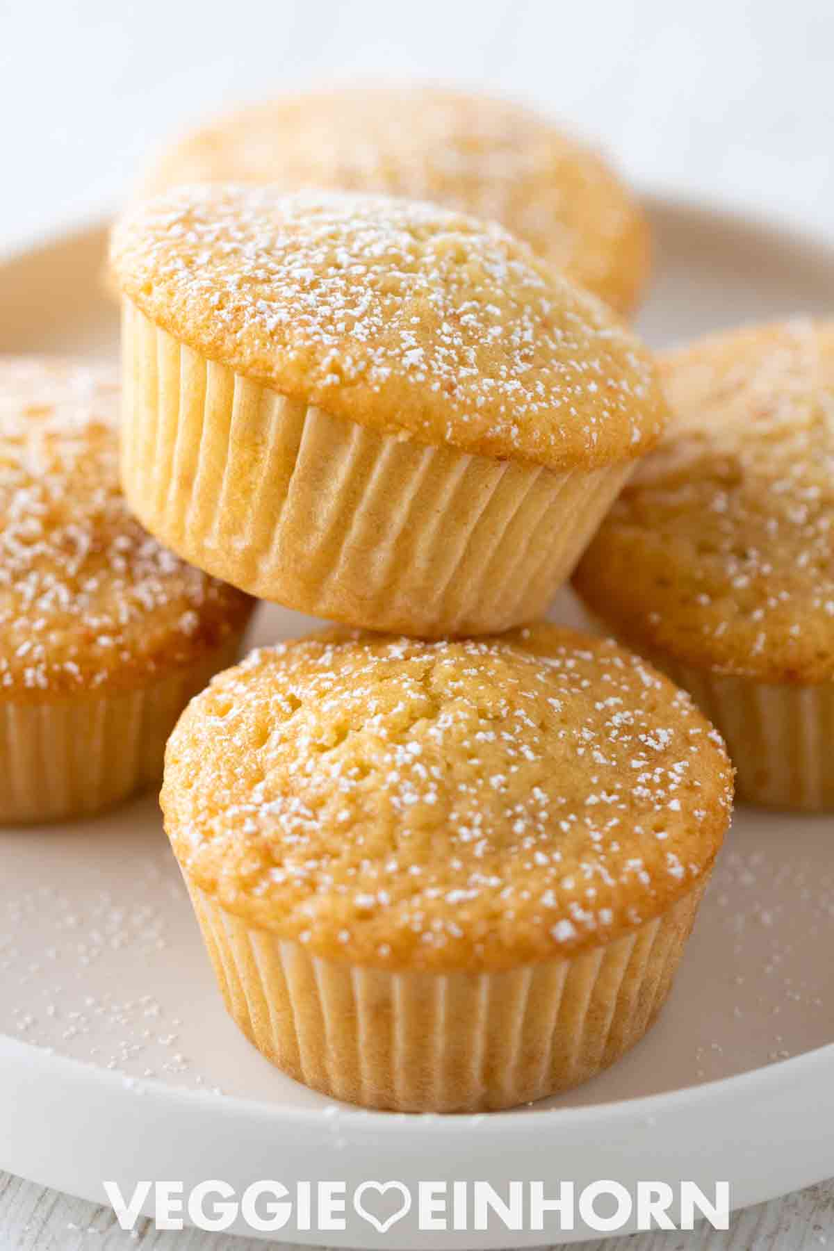 Mehrere goldbraune vegane Muffins in Papierförmchen, mit Puderzucker bestäubt, auf einem weißen Teller serviert.