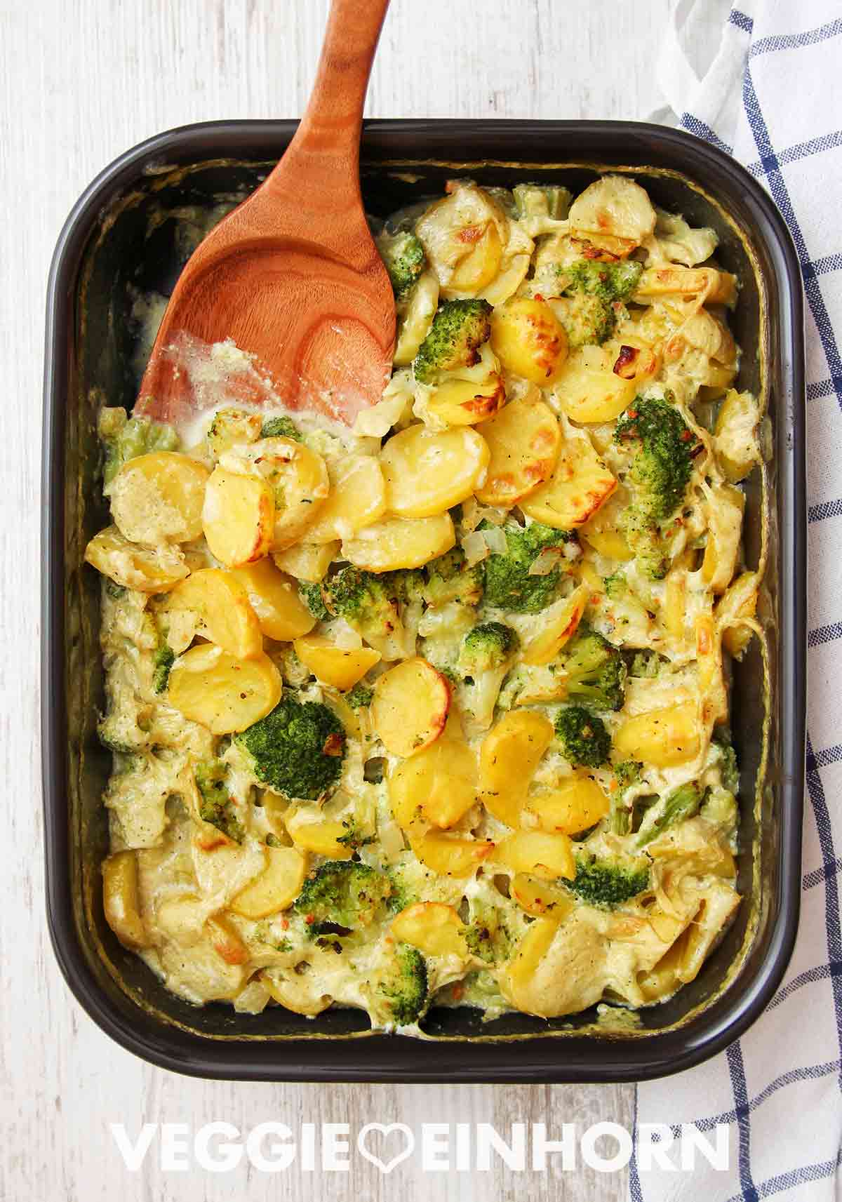 In einer Auflaufform gebackener veganer Auflauf mit Kartoffelscheiben und Brokkoli. In der Auflaufform liegt ein Holzlöffel, daneben ein blau-weiß kariertes Küchentuch.