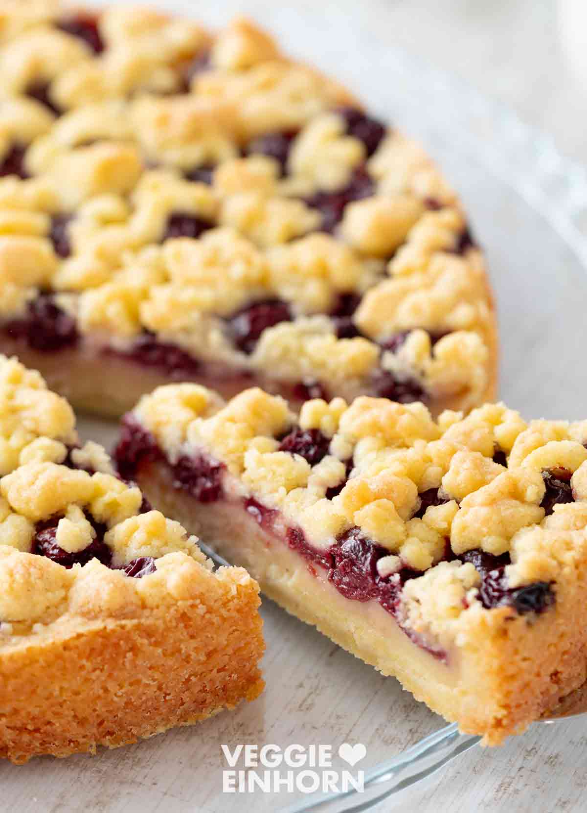 Ein veganer Kirsch Pudding Kuchen mit Streuseln in Nahaufnahme. Ein Stück wird auf einem Kuchenheber herausgehoben.