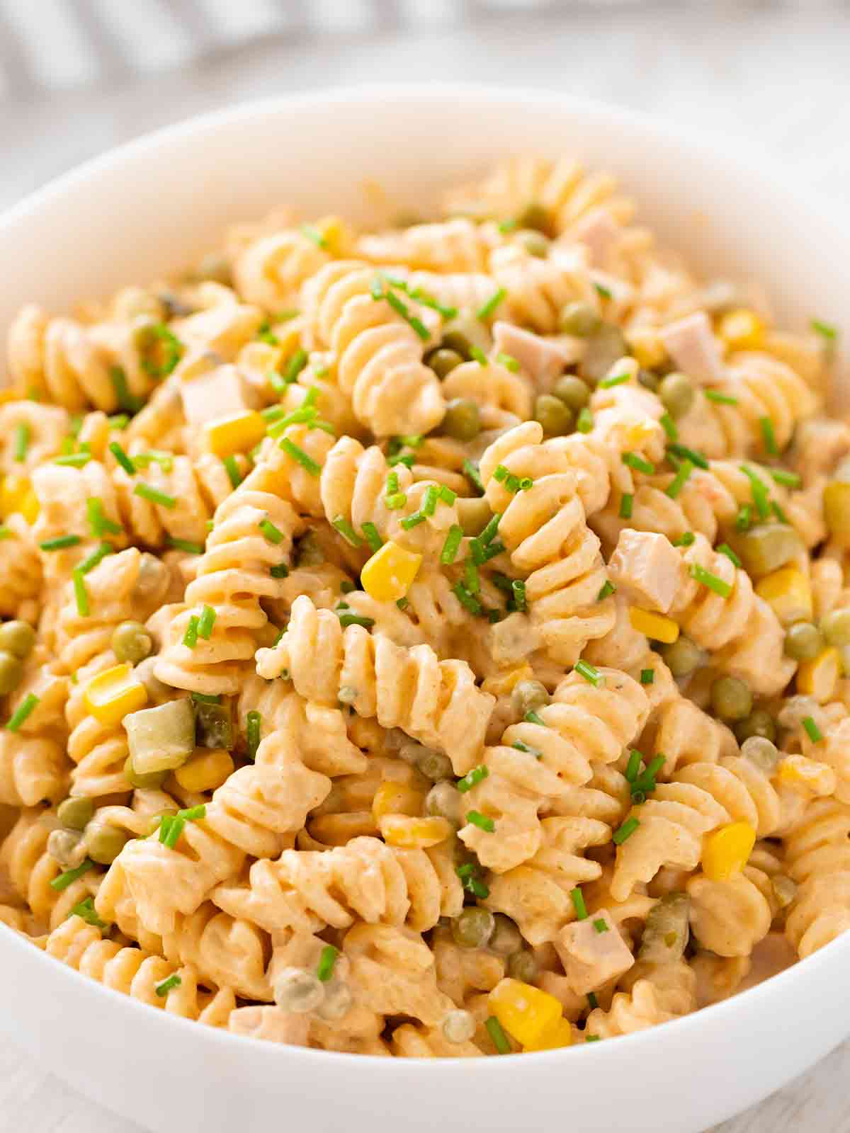 Nahaufnahme einer Schüssel mit cremigem veganem Nudelsalat aus Fusilli, veganer Fleischwurst, Erbsen, Mais und Gewürzgurken, bestreut mit frischem Schnittlauch.