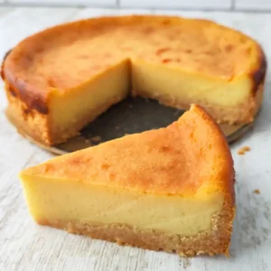 Ein Stück kleiner veganer Käsekuchen liegt vor dem angeschnittenen Kuchen auf einer weißen Holzoberfläche.