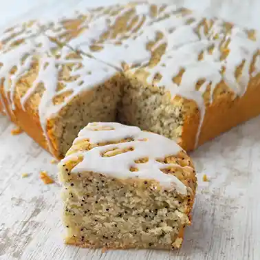 Veganer Zitronen-Mohnkuchen mit angeschnittenem Kuchenstück auf einer weißen Holzoberfläche.