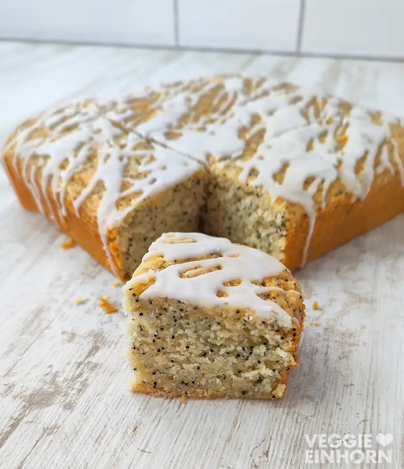 Ein veganer Mohn-Zitronenkuchen mit Zuckerguss auf einer weißen Holzoberfläche, vor dem Kuchen liegt ein angeschnittenes Stück.