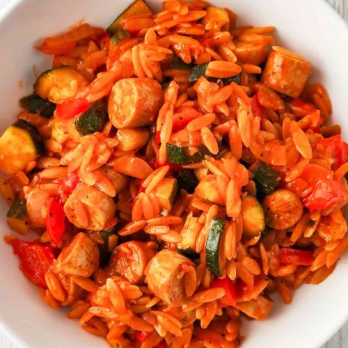 Draufsicht auf einen weißen Teller mit einer Gemüsepfanne aus Kritharaki-Nudeln, veganen Bratwurststücken, Zucchini und Paprika in einer Tomatensoße.