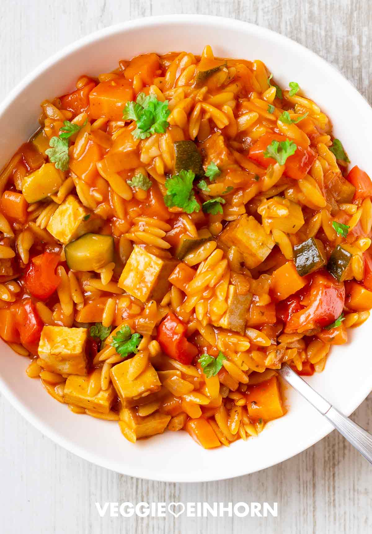 Vegane Kritharaki-Pfanne mit Räuchertofu, Zucchini, Paprika und Möhren, serviert auf einem weißen Teller und garniert mit frischer Petersilie.
