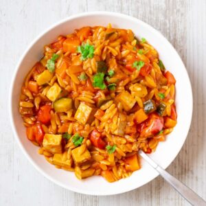 Vegane Kritharaki-Pfanne mit Räuchertofu und Gemüse in einem weißen Teller, garniert mit frischer Petersilie.
