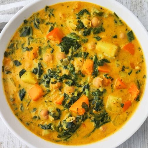 Eine Nahaufnahme zeigt einen cremigen Kürbis Kichererbsen Eintopf mit Spinat, Kartoffeln und Kürbisstücken in einer weißen Schüssel auf einer hellen Oberfläche.