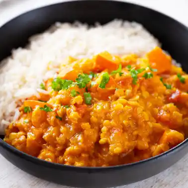 Veganes Kürbiscurry mit roten Linsen in einem schwarzen Teller, serviert mit Reis und frischer Petersilie.