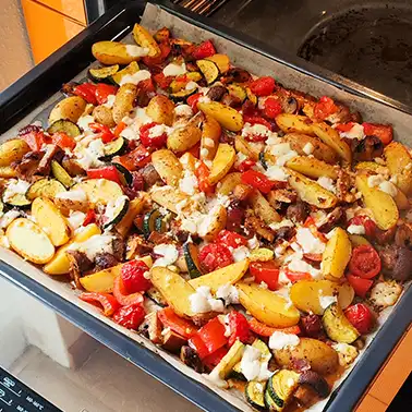 Frisch gebackenes mediterranes Ofengemüse mit Kartoffeln und veganem Feta auf einem Blech im geöffneten Backofen.