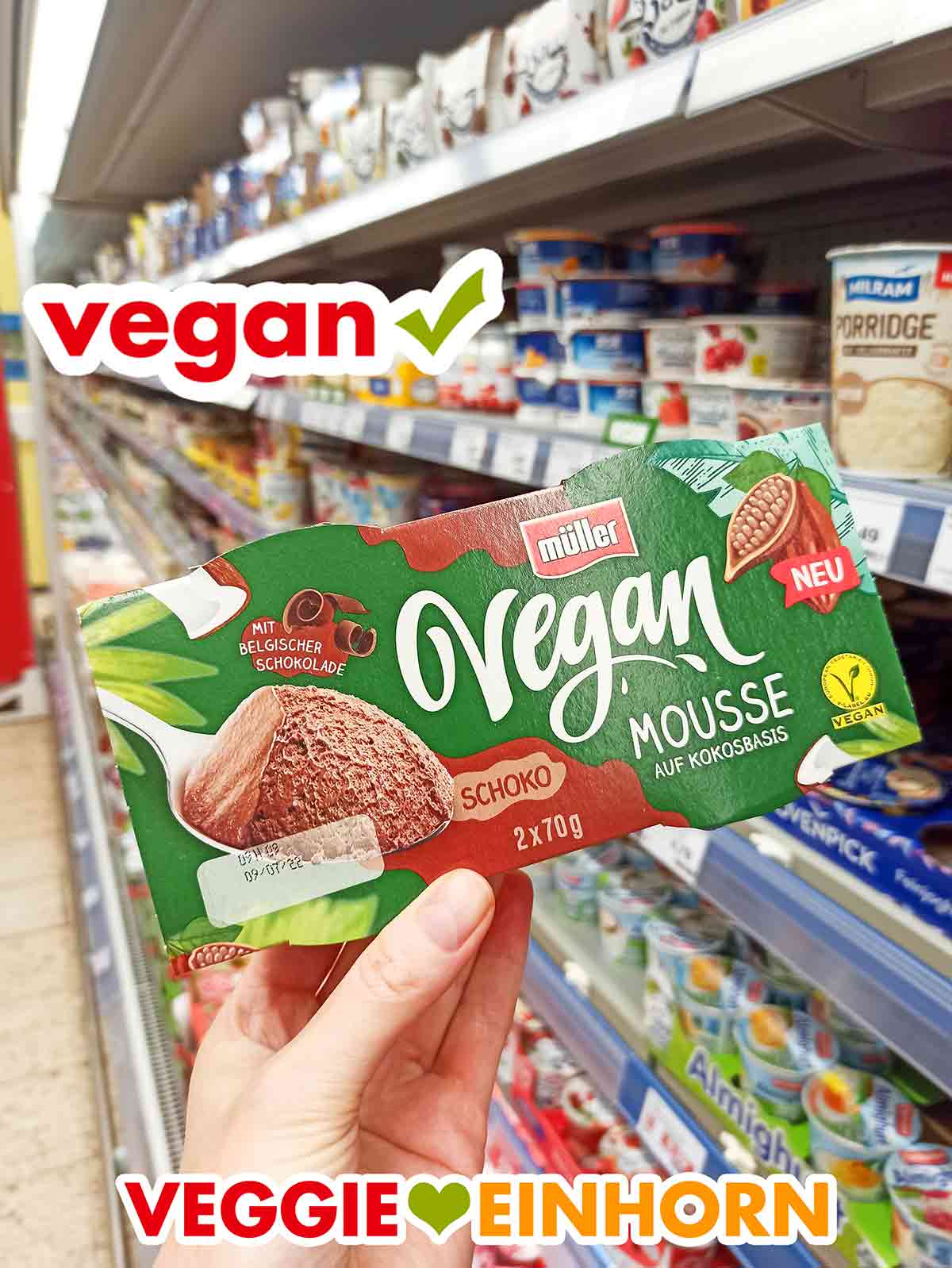 Das vegane Mousse au Chocolat von Müller Milch gibt es zum Beispiel bei Edeka. Eine Hand hält eine Packung Müller Vegan Mousse Schoko vor dem Kühlregal bei Edeka