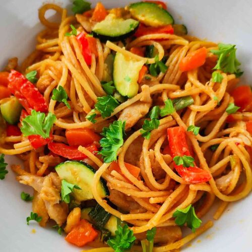 Nahaufnahme einer veganen Nudelpfanne mit Erdnusssoße, Spaghetti, roten Paprikastücken, Zucchini und veganem Hähnchen.