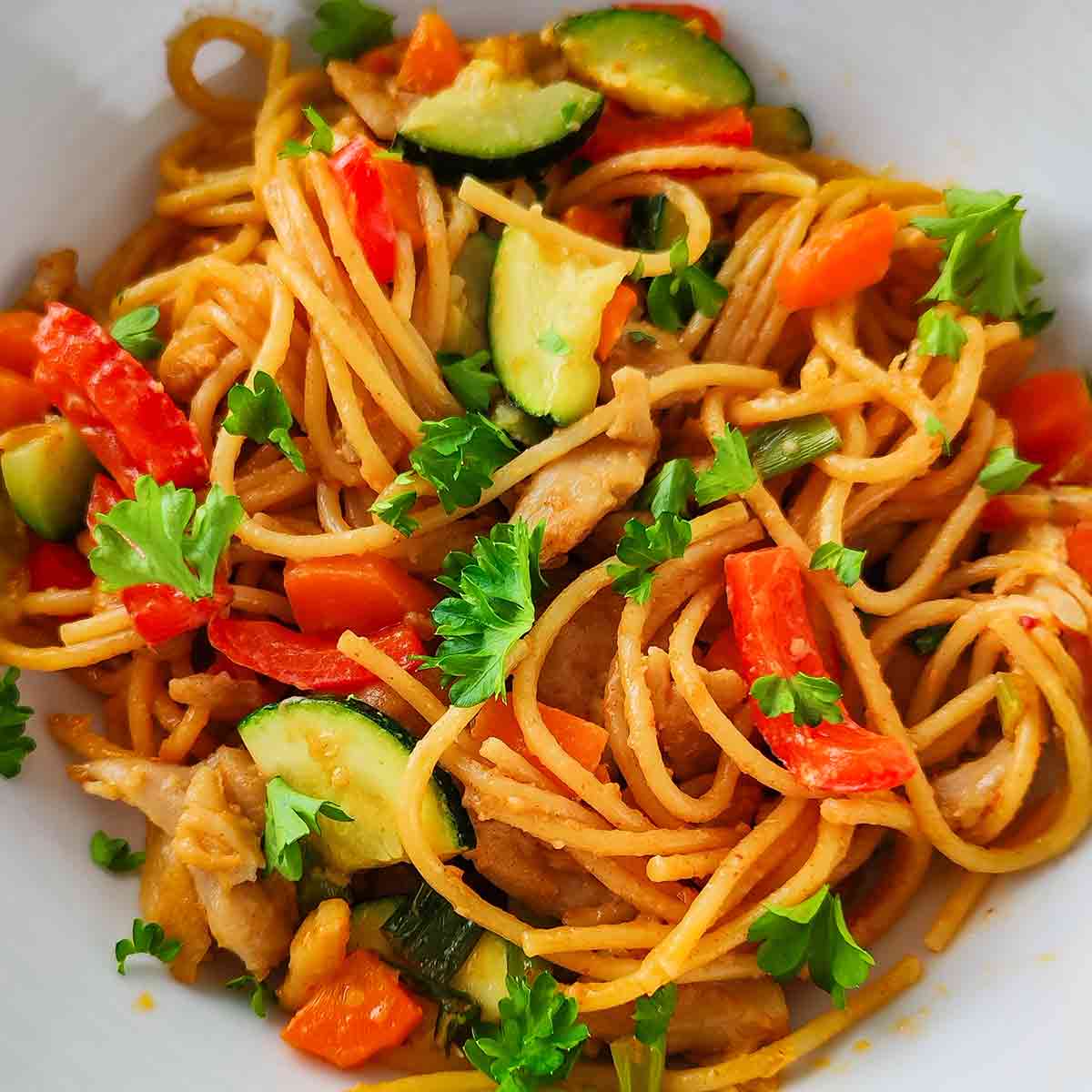 Nahaufnahme einer veganen Nudelpfanne mit Erdnusssoße, Spaghetti, roten Paprikastücken, Zucchini und veganem Hähnchen.