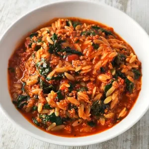 Veganer Kritharaki-Eintopf mit Blattspinat, Tomaten und veganem Hackfleisch in einem weißen Teller auf hellem Holztisch.