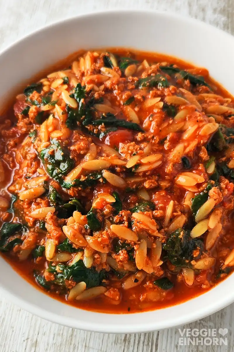 Ein weißer Teller mit veganem Spinat-Orzo-Eintopf mit veganem Hack und Tomaten.