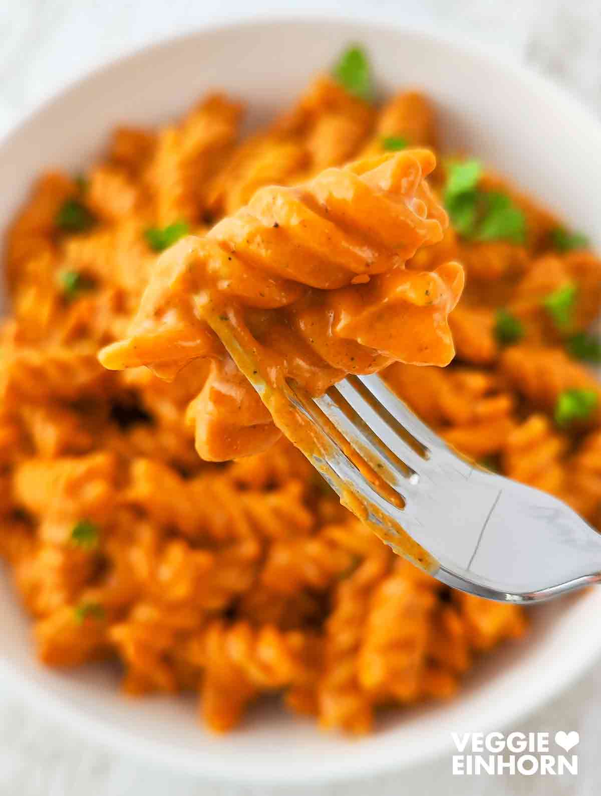 Nahaufnahme einer Gabel mit Fusilli mit orangefarbener Gemüsesoße, im Hintergrund ein weißer Teller mit weiteren Nudeln und Petersilie.