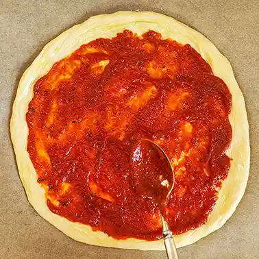 Auf einem runden Pizzateig, der auf Backpapier liegt, wird mit einem Löffel eine vegane Pizzasoße gleichmäßig verteilt.