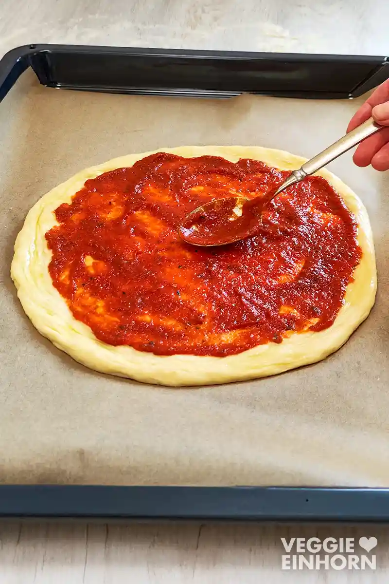 Eine Hand verteilt mit einem Löffel vegane Pizzasoße auf einem runden Pizzateig, der auf einem mit Backpapier belegten Backblech liegt.