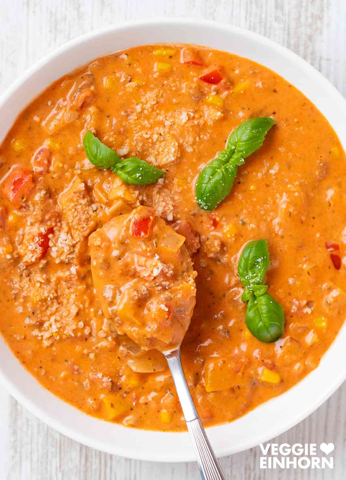 Cremige vegane Pizzasuppe mit veganem Hackfleisch, Paprika und Mais, serviert in einer weißen Schüssel. Ein Löffel mit Pizzasuppe wird aus der Schüssel gehoben. Garniert mit frischem Basilikum und veganem Käseersatz.