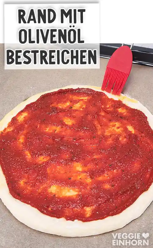 Der Rand eines mit Pizzasoße bestrichenen Pizzateiges wird mit einem roten Silikonpinsel mit Olivenöl bestrichen.