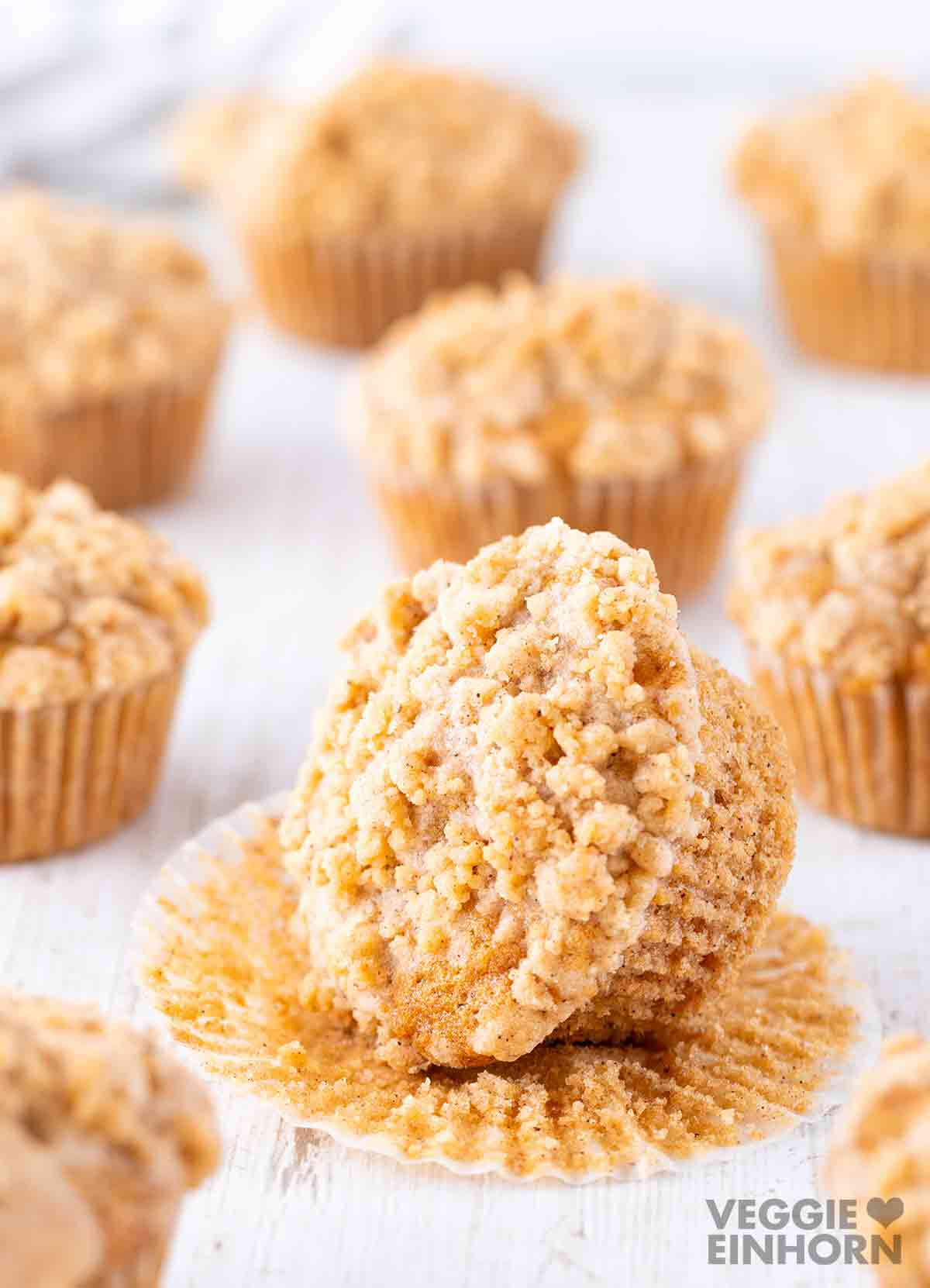 Saftiger veganer Apfelmuffin mit Streuseln, ausgepackt aus dem Papierförmchen, im Vordergrund, mit weiteren Muffins im Hintergrund.