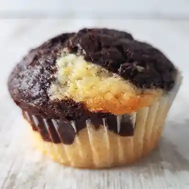 Ein einzelner veganer Marmormuffin in einer weißen Papierform auf heller Holzoberfläche mit klar sichtbarer Marmorierung aus hellem Vanilleteig und dunklem Schokoladenteig.