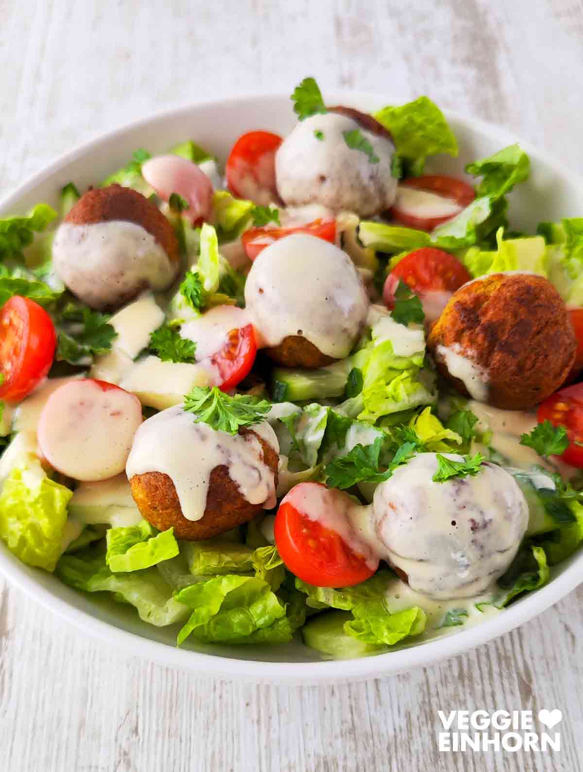 Gemischter Salat mit knusprigen Falafeln, Kirschtomaten, Gurken und cremigem Dressing, serviert in einer weißen Schüssel auf hellem Holztisch.