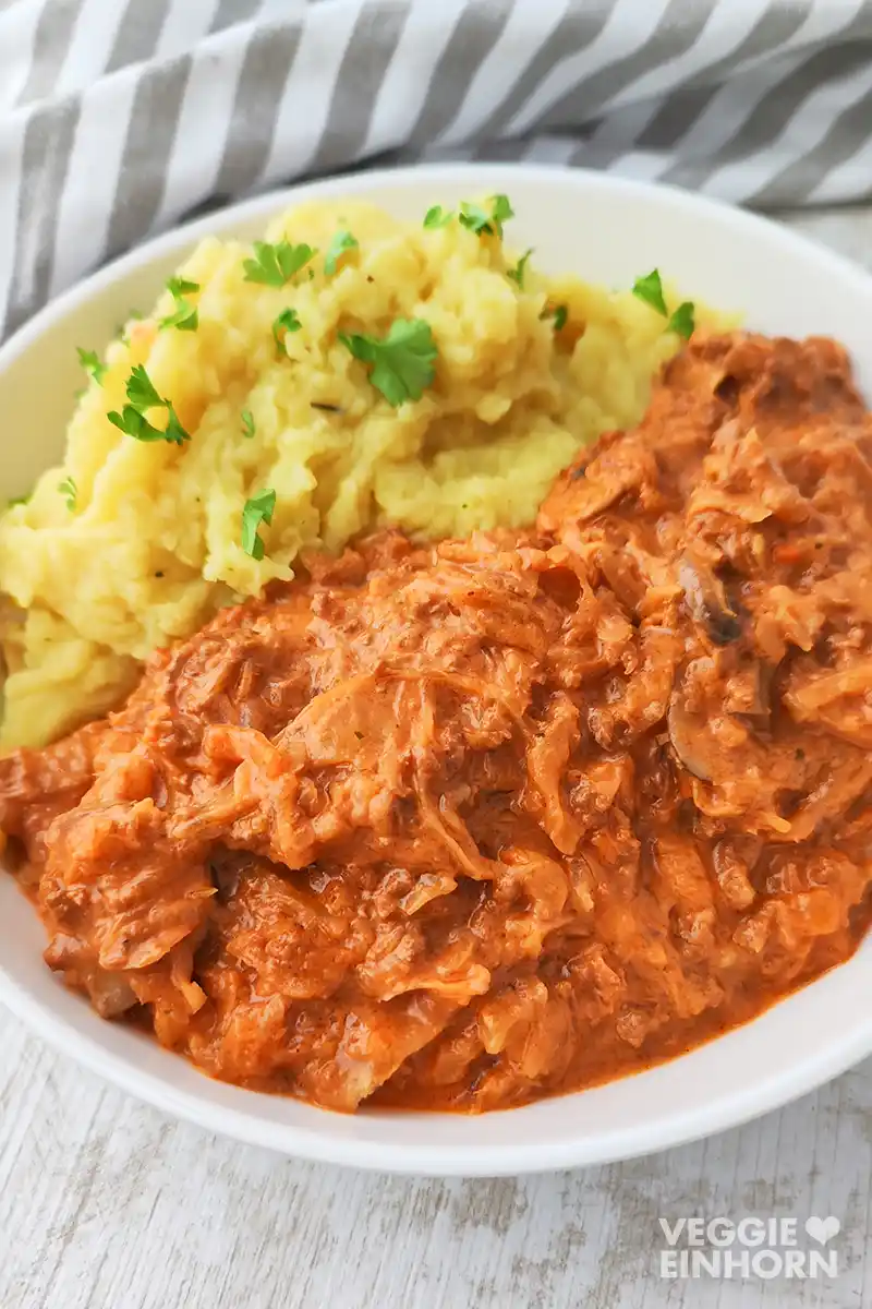 Sauerkrautpfanne mit veganem Hack und Champignons Nahaufnahme einer Portion veganer Sauerkrautpfanne und Kartoffelpüree auf einem weißen Teller.