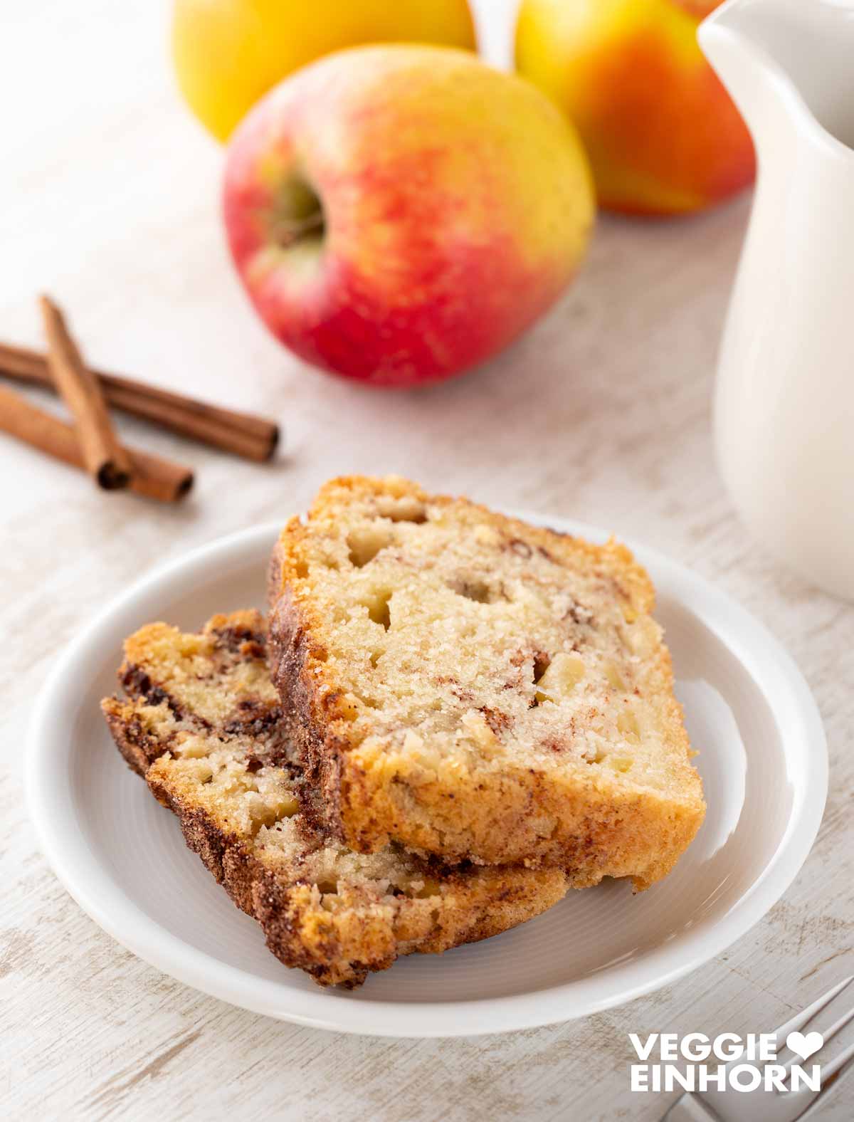 Zwei Stücke veganer Zimtkuchen mit Apfel auf einem kleinen weißen Teller, umgeben von frischen Äpfeln, Zimtstangen und einer kleinen weißen Kanne im Hintergrund.