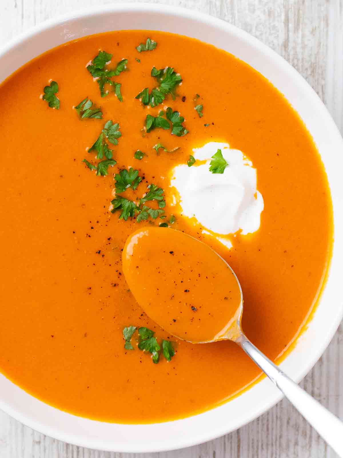 Weißer Teller mit Tomatensuppe, dekoriert mit Petersilie und einem Klecks Kokosmilch. Ein Löffel mit Suppe liegt im Teller.