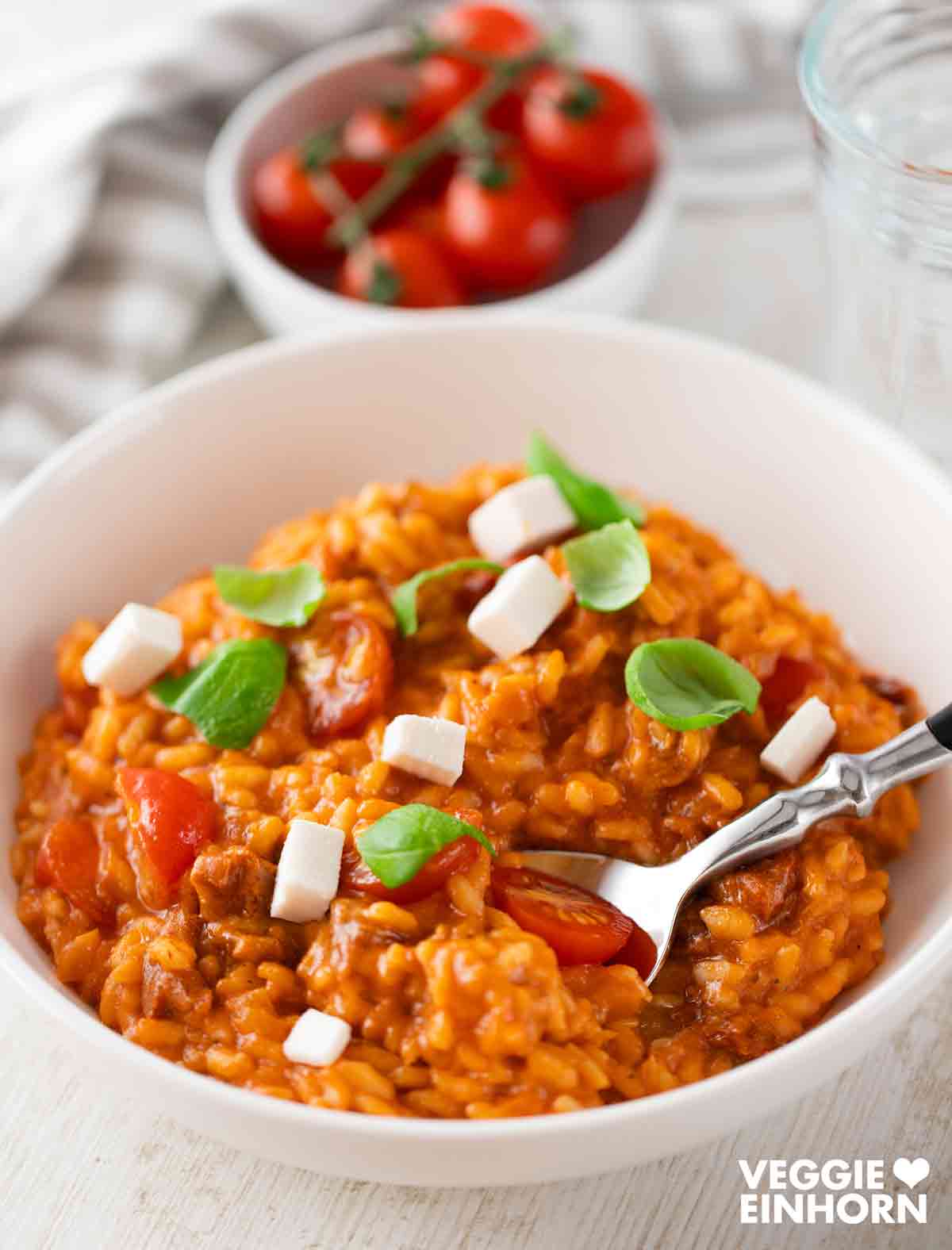 Veganes Tomatenrisotto, garniert mit Basilikum, Kirschtomaten und veganen Fetawürfeln, serviert in einer weißen Schale.