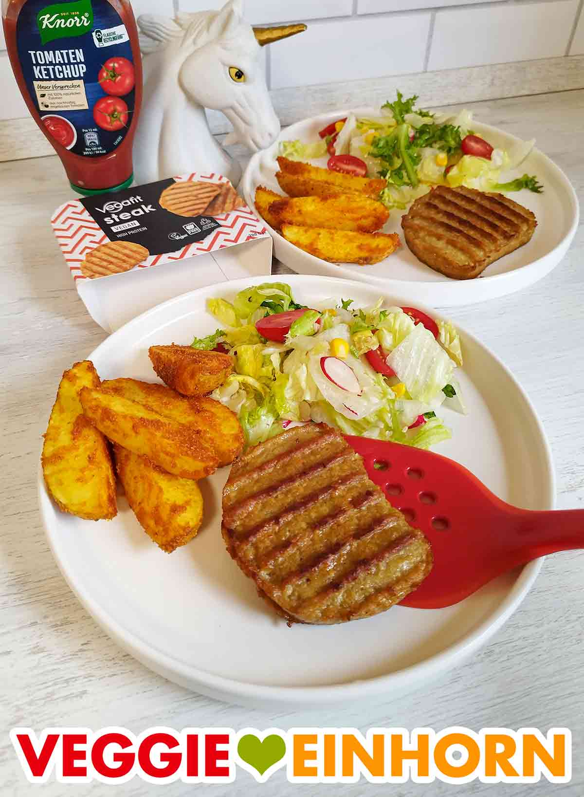 Die Vegafit Steaks passen gut zu panierten Kartoffeln und einem frischen Salat. Zwei Teller mit veganen Steaks, panierten Kartoffelspalten und Salat