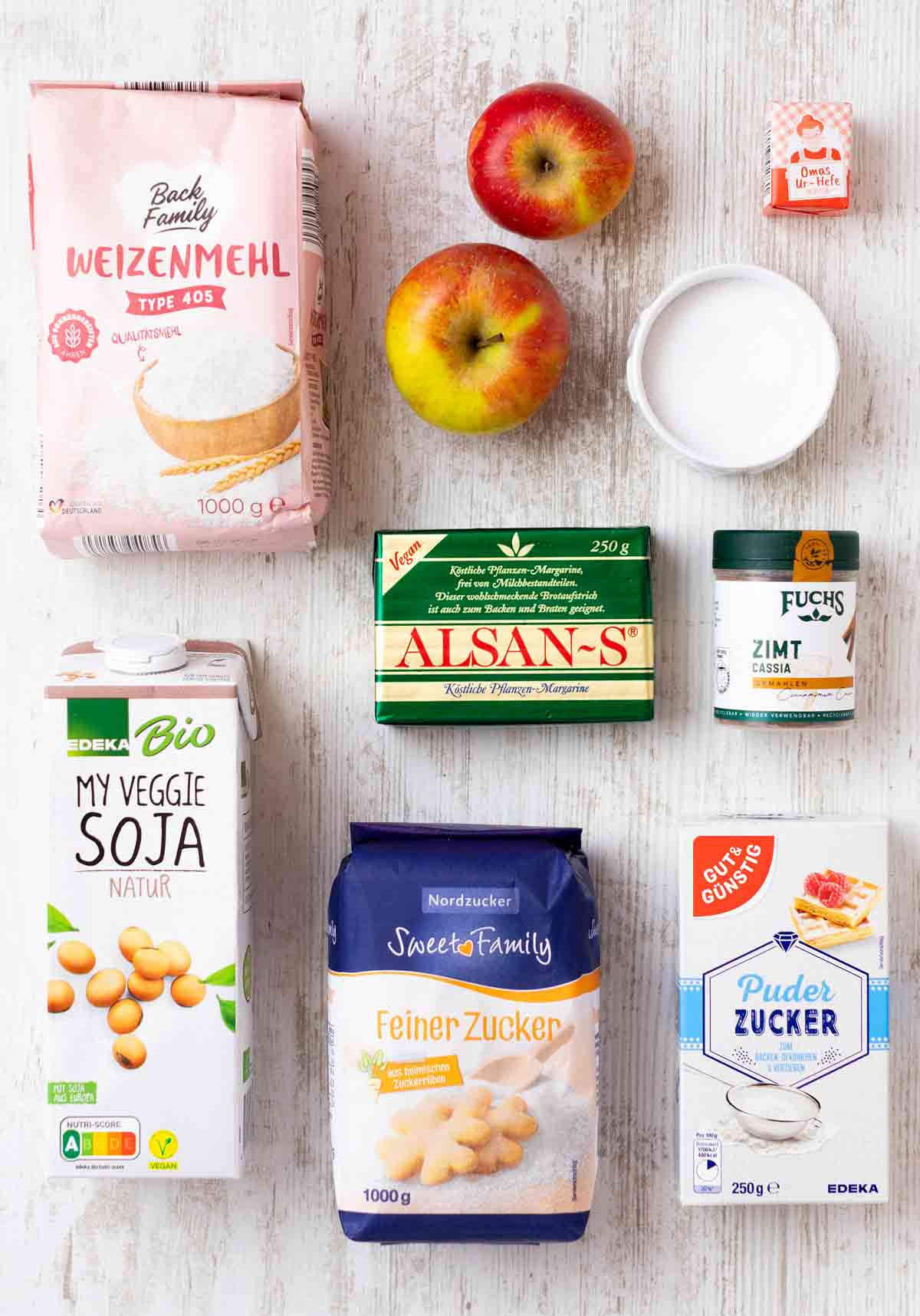 Auf einem weißen Holzuntergrund liegen Zutaten für vegane Apfel-Zimt-Schnecken: Weizenmehl, zwei Äpfel, ein Würfel Hefe, eine Salzschale, Zimt, vegane Butter, Sojamilch, Zucker und Puderzucker.