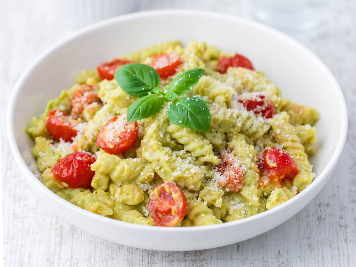Vegane Avocado Pasta mit Kirschtomaten und Basilikum auf einem weißen Teller, bestreut mit veganem Parmesan.