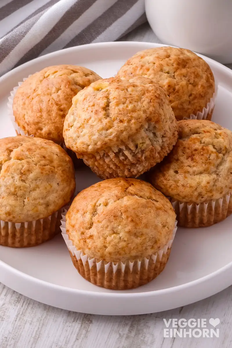 Vegane Bananenbrot Muffins auf einem weißen Teller neben einem gestreiften Küchentuch.