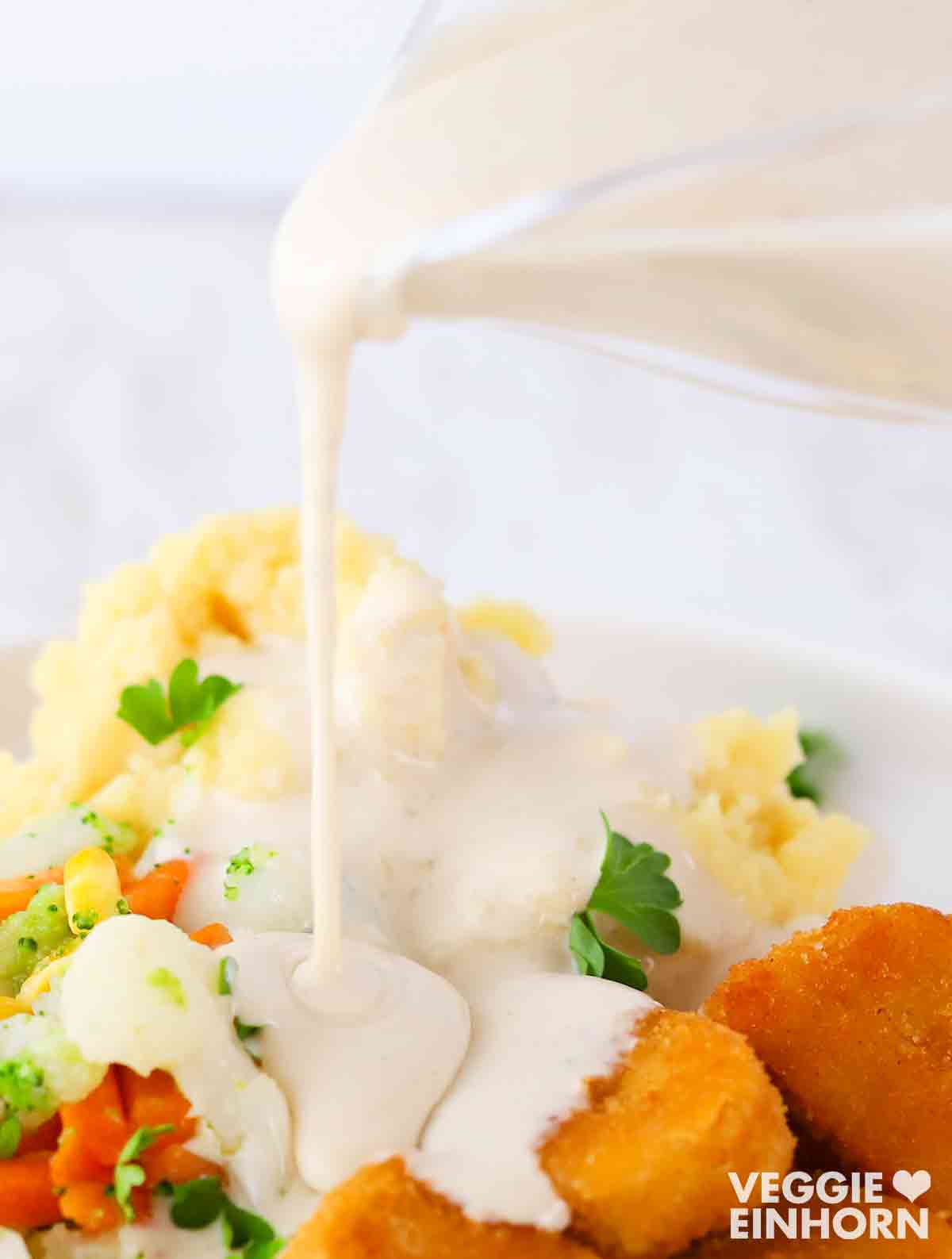 Vegane Béchamelsauce wird über Kartoffelpüree, Gemüse und vegane Nuggets auf einem Teller gegossen.