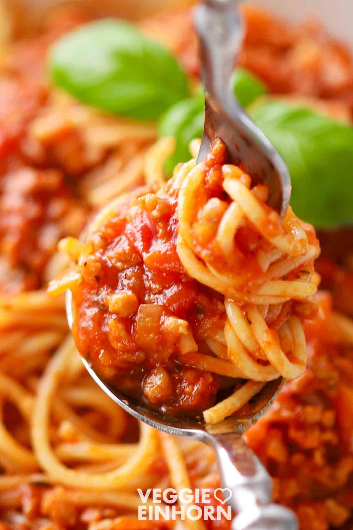 Eine Nahaufnahme von veganer Bolognese mit Sojagranulat und Spaghetti, eingerollt auf einer Gabel, garniert mit frischem Basilikum im Hintergrund.