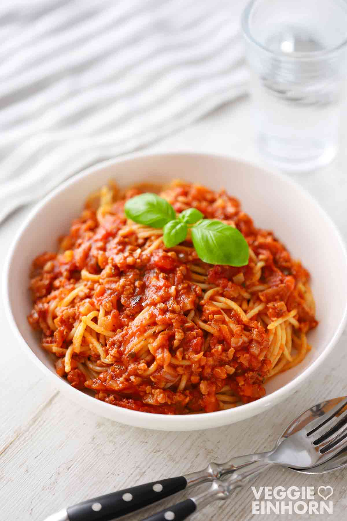 Ein Teller vegane Bolognese mit Sojagranulat und Spaghetti, garniert mit frischem Basilikum, mit Besteck und einem Glas Wasser im Hintergrund.