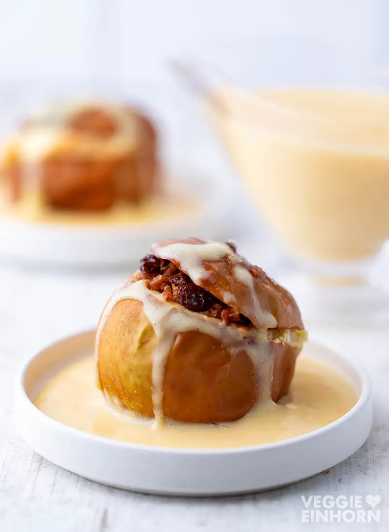 Veganer Bratapfel mit Spekulatius und Rosinen Ein Bratapfel mit veganer Vanillesoße und Rosinenfüllung auf einem weißen Teller, im Hintergrund unscharf eine Sauciere.