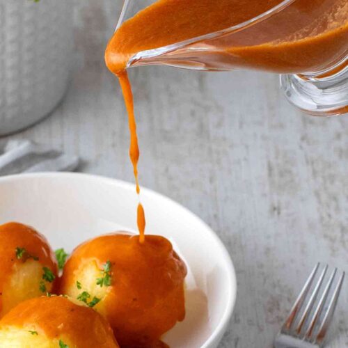 Vegane braune Soße wird aus einer Sauciere über drei Kartoffelklöße in einem weißen Teller gegossen, garniert mit frischer Petersilie.