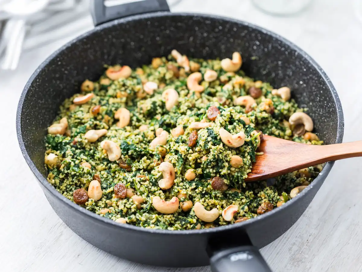 Vegane Couscous Pfanne mit Kichererbsen, Spinat, Cashews und Rosinen in einer schwarzen Pfanne mit einem Holzlöffel.